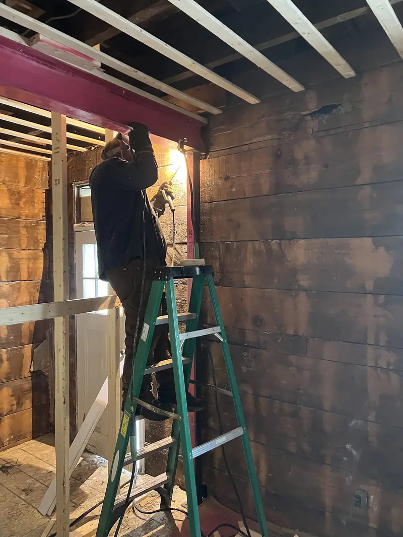 A welder on a green stepladder works on a red steel beam under wooden joists in a gutted old room
