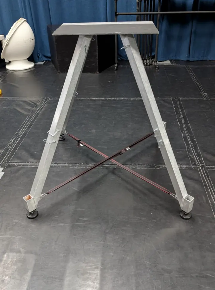 A tall A-frame metal stand with square-tube legs, crossed braces and a flat top plate, standing on a dark floor