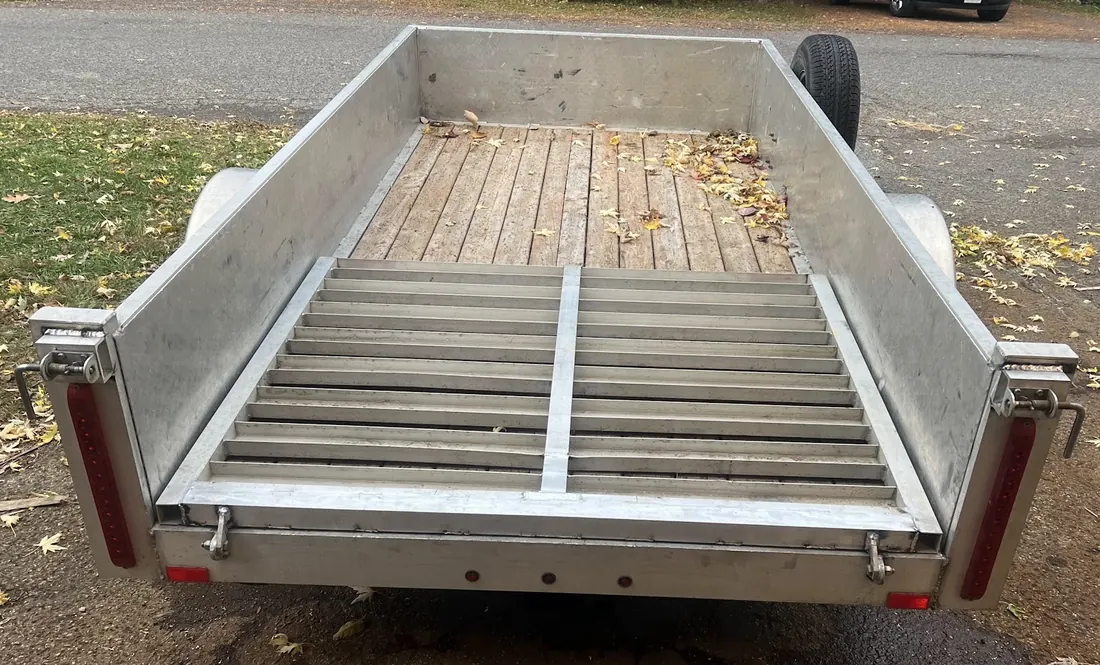 Rear of an aluminum utility trailer with a wooden deck and the ramp folded up, parked on a leafy street